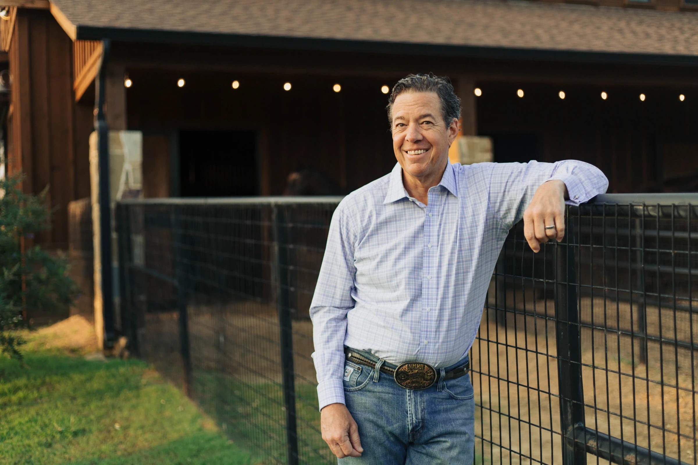 A lawyer leans against a wooden rail outside a ranch barn in warm evening light.